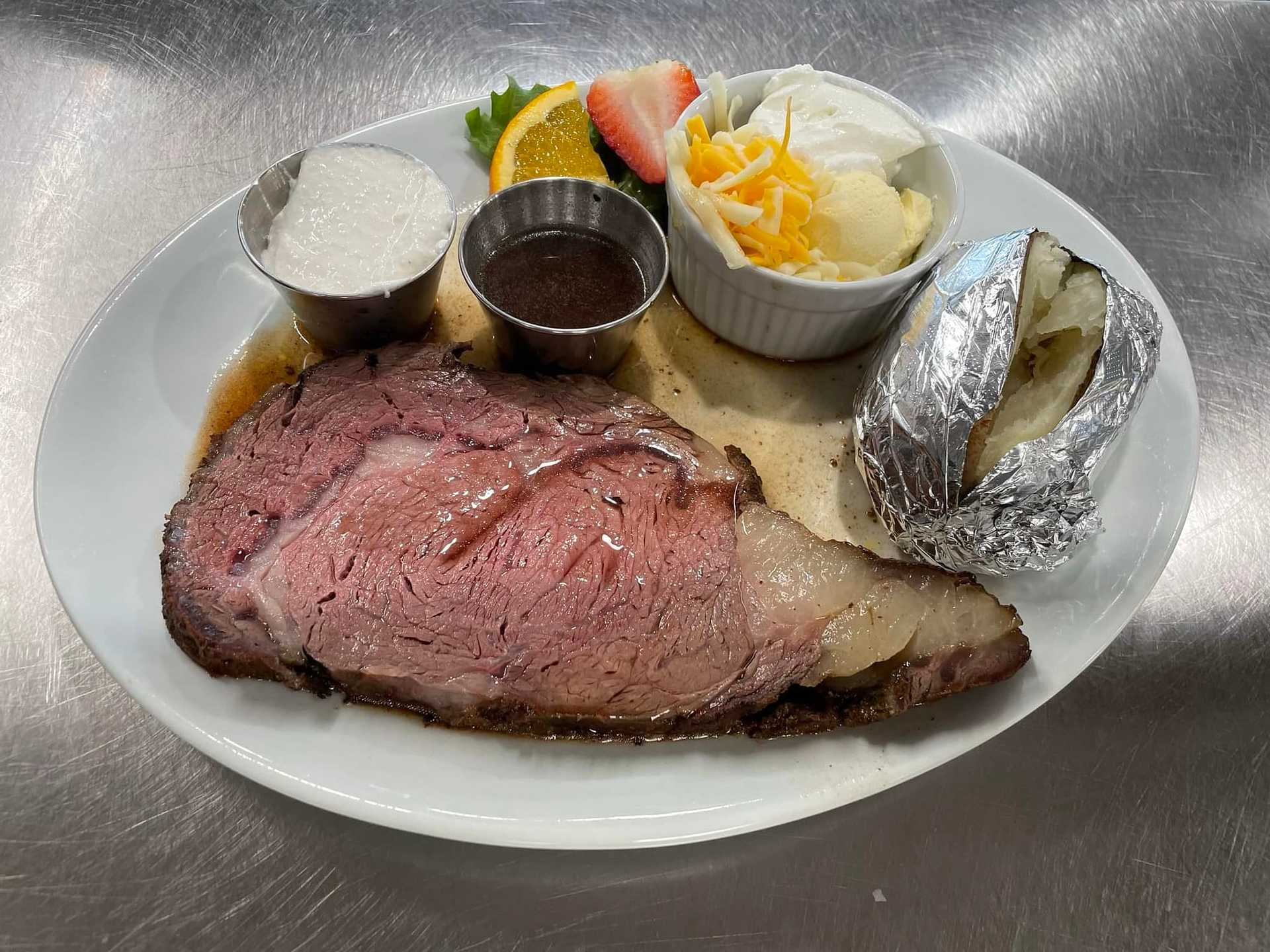 Plate of prime rib with baked potato, sauces, cheese, fruit garnish, and a buttered bread slice.