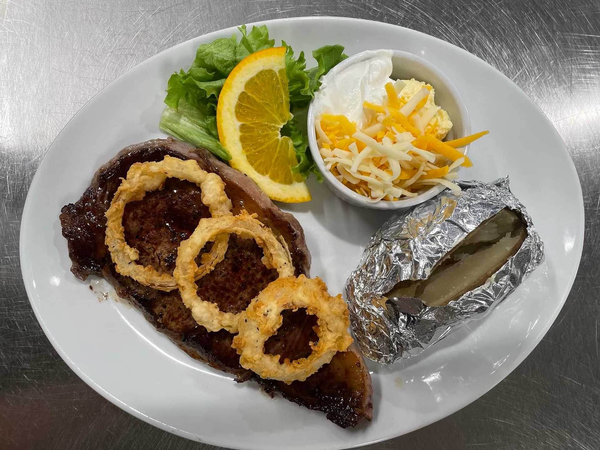 Steak with onion rings, baked potato, salad, sour cream, and cheese on a white plate.