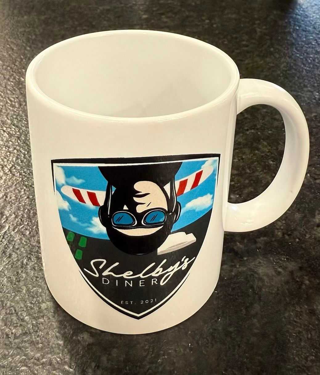 White mug with "Shelby's Diner" logo featuring a pilot and plane, established in 2021.