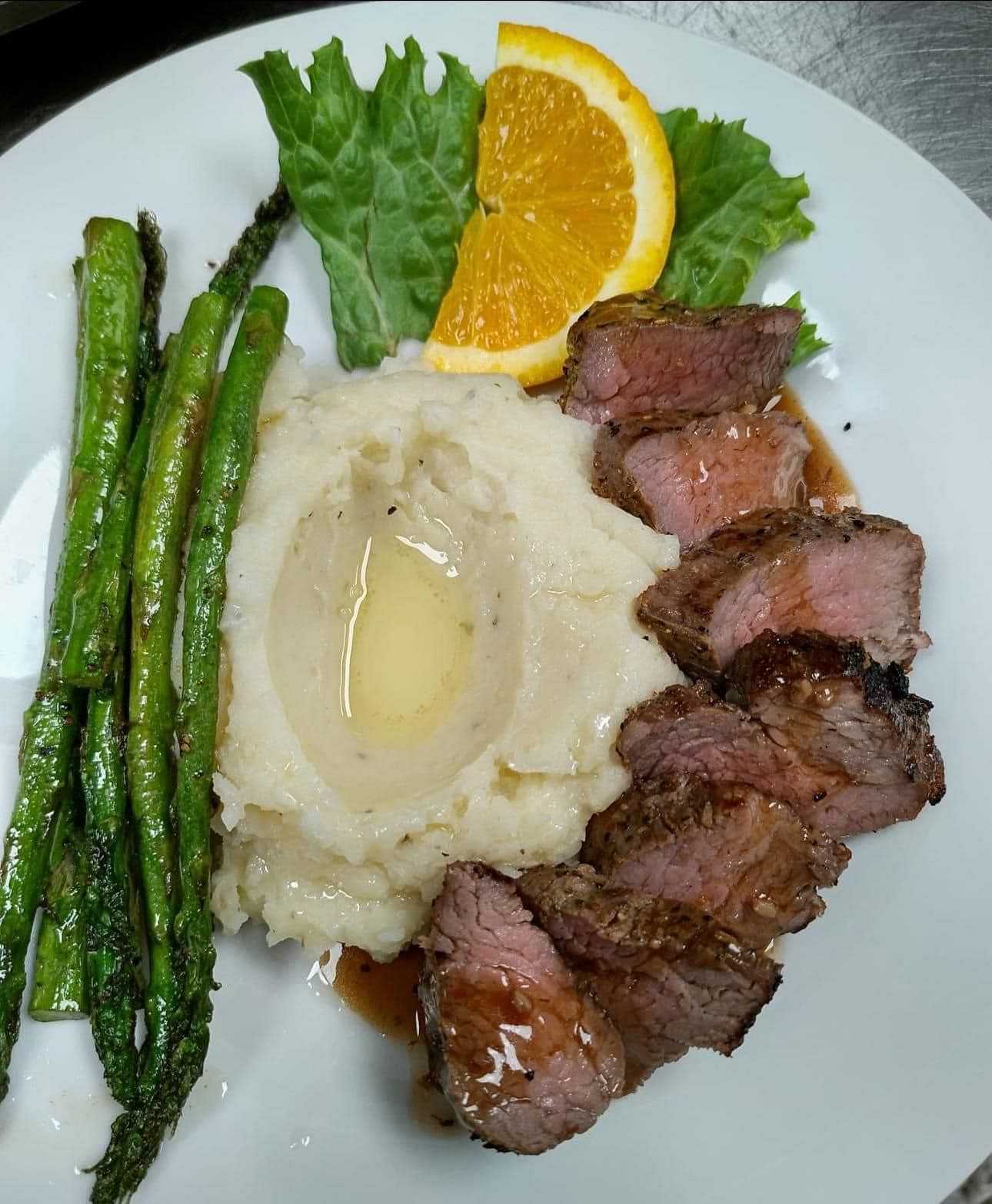 Sliced steak, mashed potatoes, asparagus, and an orange slice on a plate with a lettuce garnish.